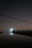 A large satellite dish is illuminated against a dark night sky, reflecting in a calm body of water in the foreground. The dish is surrounded by silhouetted trees and structures with faint, distant city lights in the background. Overhead, power lines stretch across the scene.