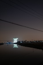 A large satellite dish is illuminated against a dark night sky, reflecting in a calm body of water in the foreground. The dish is surrounded by silhouetted trees and structures with faint, distant city lights in the background. Overhead, power lines stretch across the scene.