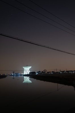 A large satellite dish is illuminated against a dark night sky, reflecting in a calm body of water in the foreground. The dish is surrounded by silhouetted trees and structures with faint, distant city lights in the background. Overhead, power lines stretch across the scene.