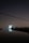 A large satellite dish is illuminated against a dark night sky, reflecting in a calm body of water in the foreground. The dish is surrounded by silhouetted trees and structures with faint, distant city lights in the background. Overhead, power lines stretch across the scene.