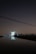 A large satellite dish is illuminated against a dark night sky, reflecting in a calm body of water in the foreground. The dish is surrounded by silhouetted trees and structures with faint, distant city lights in the background. Overhead, power lines stretch across the scene.