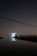 A large satellite dish is illuminated against a dark night sky, reflecting in a calm body of water in the foreground. The dish is surrounded by silhouetted trees and structures with faint, distant city lights in the background. Overhead, power lines stretch across the scene.