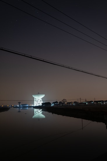 A large satellite dish is illuminated against a dark night sky, reflecting in a calm body of water in the foreground. The dish is surrounded by silhouetted trees and structures with faint, distant city lights in the background. Overhead, power lines stretch across the scene.