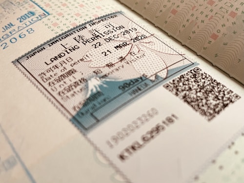 A close-up view of a Japanese immigration inspector's landing permission stamp on a passport page, featuring a document with text in both Japanese and English. The stamp includes the date of entry and validity, with visible QR codes and security features embedded in the design.
