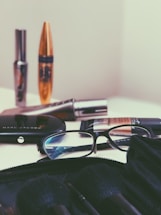 A collection of cosmetic items is spread out on a surface, including mascara tubes, an eyeshadow palette, and a foundation. A pair of reading glasses rests among the items, positioned next to the palette. The background and overall setting seem minimalistic, focusing mainly on the makeup products.