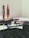 A collection of cosmetic items is spread out on a surface, including mascara tubes, an eyeshadow palette, and a foundation. A pair of reading glasses rests among the items, positioned next to the palette. The background and overall setting seem minimalistic, focusing mainly on the makeup products.