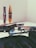 A collection of cosmetic items is spread out on a surface, including mascara tubes, an eyeshadow palette, and a foundation. A pair of reading glasses rests among the items, positioned next to the palette. The background and overall setting seem minimalistic, focusing mainly on the makeup products.