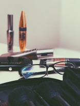 A collection of cosmetic items is spread out on a surface, including mascara tubes, an eyeshadow palette, and a foundation. A pair of reading glasses rests among the items, positioned next to the palette. The background and overall setting seem minimalistic, focusing mainly on the makeup products.