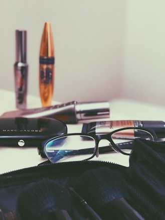 A collection of cosmetic items is spread out on a surface, including mascara tubes, an eyeshadow palette, and a foundation. A pair of reading glasses rests among the items, positioned next to the palette. The background and overall setting seem minimalistic, focusing mainly on the makeup products.