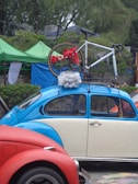 A vintage blue and cream Volkswagen Beetle is parked with a bicycle mounted on its roof rack. The bicycle is decorated with red flowers and white fluffy material. The scene is set outdoors with green tents and trees in the background, adding a natural element to the setting. A red car is partially visible in the foreground.