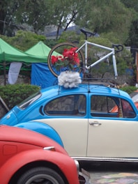 A vintage blue and cream Volkswagen Beetle is parked with a bicycle mounted on its roof rack. The bicycle is decorated with red flowers and white fluffy material. The scene is set outdoors with green tents and trees in the background, adding a natural element to the setting. A red car is partially visible in the foreground.