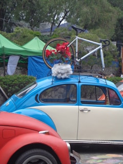 A vintage blue and cream Volkswagen Beetle is parked with a bicycle mounted on its roof rack. The bicycle is decorated with red flowers and white fluffy material. The scene is set outdoors with green tents and trees in the background, adding a natural element to the setting. A red car is partially visible in the foreground.