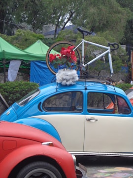 A vintage blue and cream Volkswagen Beetle is parked with a bicycle mounted on its roof rack. The bicycle is decorated with red flowers and white fluffy material. The scene is set outdoors with green tents and trees in the background, adding a natural element to the setting. A red car is partially visible in the foreground.