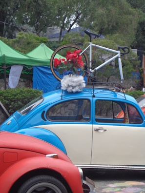 A vintage blue and cream Volkswagen Beetle is parked with a bicycle mounted on its roof rack. The bicycle is decorated with red flowers and white fluffy material. The scene is set outdoors with green tents and trees in the background, adding a natural element to the setting. A red car is partially visible in the foreground.
