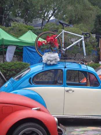 A vintage blue and cream Volkswagen Beetle is parked with a bicycle mounted on its roof rack. The bicycle is decorated with red flowers and white fluffy material. The scene is set outdoors with green tents and trees in the background, adding a natural element to the setting. A red car is partially visible in the foreground.