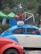 A vintage blue and cream Volkswagen Beetle is parked with a bicycle mounted on its roof rack. The bicycle is decorated with red flowers and white fluffy material. The scene is set outdoors with green tents and trees in the background, adding a natural element to the setting. A red car is partially visible in the foreground.