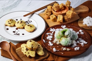 This is an arrangement of various traditional snacks and sweets displayed on plates and wooden boards. There are round fritters with chocolate sprinkles, green coconut-covered balls, crunchy fried cubes, and baked goods with a cheese-like filling. Wooden utensils and a small pile of shredded coconut are also on the table.