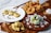 This is an arrangement of various traditional snacks and sweets displayed on plates and wooden boards. There are round fritters with chocolate sprinkles, green coconut-covered balls, crunchy fried cubes, and baked goods with a cheese-like filling. Wooden utensils and a small pile of shredded coconut are also on the table.