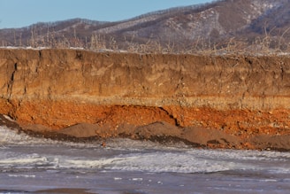 A soil profile is exposed with distinct layers of earth showing variations in color and texture. The top layer is dark brown, transitioning to a reddish-brown layer below, followed by darker strata. The scene includes wintry hills in the background and patches of ice or snow at the base.