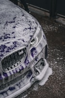 A close-up view of a car being washed, with a significant amount of white soap suds covering its purple exterior. The focus is on the front part of the vehicle, including the headlights and grille. The surrounding surface appears wet, consistent with the washing process.