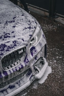 A close-up view of a car being washed, with a significant amount of white soap suds covering its purple exterior. The focus is on the front part of the vehicle, including the headlights and grille. The surrounding surface appears wet, consistent with the washing process.