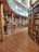 A spacious library with wooden floors and shelves, filled with a diverse collection of books. The architecture includes curved shelves and grand columns, providing an elegant and scholarly atmosphere. A glass railing separates sections of the library, and there is a computer or device stand placed near the shelves.