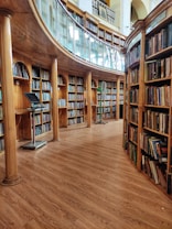 A spacious library with wooden floors and shelves, filled with a diverse collection of books. The architecture includes curved shelves and grand columns, providing an elegant and scholarly atmosphere. A glass railing separates sections of the library, and there is a computer or device stand placed near the shelves.