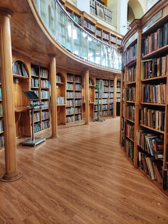 A spacious library with wooden floors and shelves, filled with a diverse collection of books. The architecture includes curved shelves and grand columns, providing an elegant and scholarly atmosphere. A glass railing separates sections of the library, and there is a computer or device stand placed near the shelves.