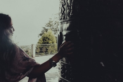A person with long hair gently touches a large, intricately carved drum or bell. The setting appears to be outdoors, near a metal fence with foliage visible in the background. The lighting is soft, creating a tranquil atmosphere.