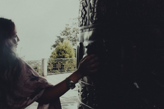 A person with long hair gently touches a large, intricately carved drum or bell. The setting appears to be outdoors, near a metal fence with foliage visible in the background. The lighting is soft, creating a tranquil atmosphere.