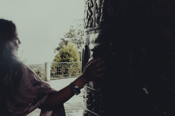 A person with long hair gently touches a large, intricately carved drum or bell. The setting appears to be outdoors, near a metal fence with foliage visible in the background. The lighting is soft, creating a tranquil atmosphere.