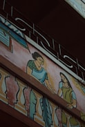 A mural painting on the exterior wall of a building depicting two women in traditional clothing. The women are illustrated with one wearing a green blouse and blue saree, and the other in a green dress, interacting on a balcony adorned with colorful decorative patterns. Above the artwork, an ornate metal railing is visible.