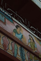 A mural painting on the exterior wall of a building depicting two women in traditional clothing. The women are illustrated with one wearing a green blouse and blue saree, and the other in a green dress, interacting on a balcony adorned with colorful decorative patterns. Above the artwork, an ornate metal railing is visible.