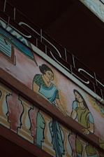 A mural painting on the exterior wall of a building depicting two women in traditional clothing. The women are illustrated with one wearing a green blouse and blue saree, and the other in a green dress, interacting on a balcony adorned with colorful decorative patterns. Above the artwork, an ornate metal railing is visible.