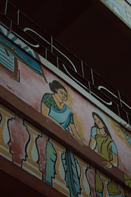 A mural painting on the exterior wall of a building depicting two women in traditional clothing. The women are illustrated with one wearing a green blouse and blue saree, and the other in a green dress, interacting on a balcony adorned with colorful decorative patterns. Above the artwork, an ornate metal railing is visible.