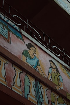 A mural painting on the exterior wall of a building depicting two women in traditional clothing. The women are illustrated with one wearing a green blouse and blue saree, and the other in a green dress, interacting on a balcony adorned with colorful decorative patterns. Above the artwork, an ornate metal railing is visible.