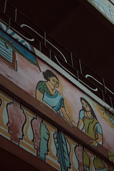 A mural painting on the exterior wall of a building depicting two women in traditional clothing. The women are illustrated with one wearing a green blouse and blue saree, and the other in a green dress, interacting on a balcony adorned with colorful decorative patterns. Above the artwork, an ornate metal railing is visible.