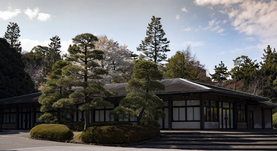 The image depicts a traditional Japanese house with a sloping roof and a large porch, surrounded by lush greenery. The house is nestled amidst trees, creating a serene atmosphere. The perspective of the photo is from street level, looking up at the house, which enhances its grandeur. The colors in the image are predominantly green and brown, reflecting the natural surroundings and architectural elements of the house.