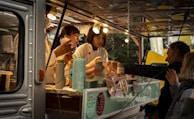 A food truck scene with two workers preparing food and interacting with customers. The workers are wearing light-colored uniforms and are busy handling utensils and food containers. Several lightbulbs illuminate the interior, and there are multiple containers and a menu visible.