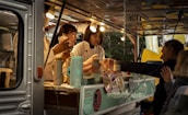 A food truck scene with two workers preparing food and interacting with customers. The workers are wearing light-colored uniforms and are busy handling utensils and food containers. Several lightbulbs illuminate the interior, and there are multiple containers and a menu visible.