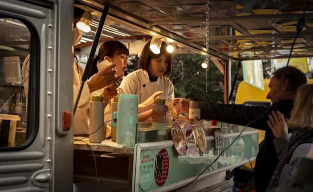 A food truck scene with two workers preparing food and interacting with customers. The workers are wearing light-colored uniforms and are busy handling utensils and food containers. Several lightbulbs illuminate the interior, and there are multiple containers and a menu visible.