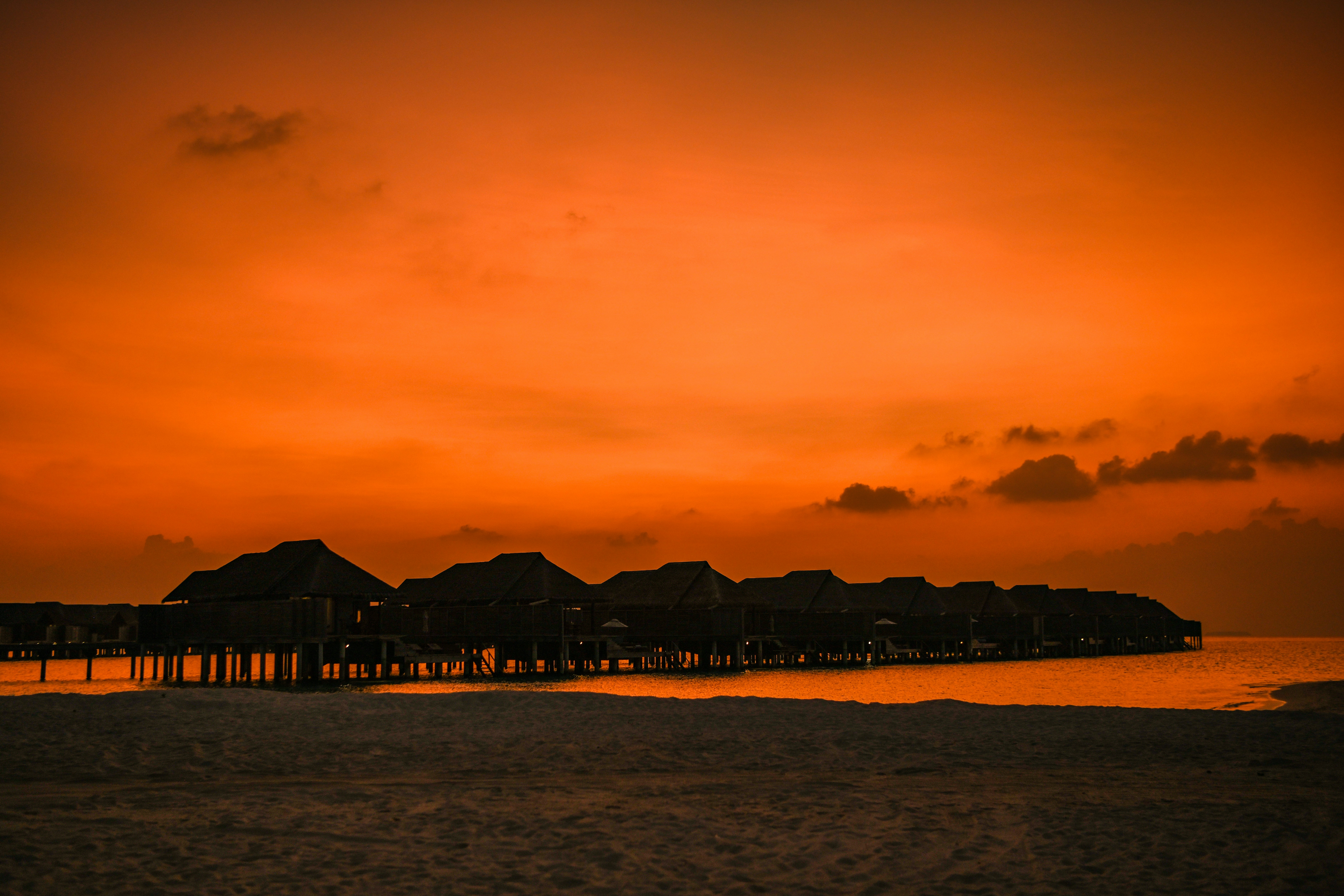 Maldives overwater bungalow deck at golden sunset, deep amber reflections on calm turquoise water, dark silhouetted palm fronds, warm luxury atmosphere