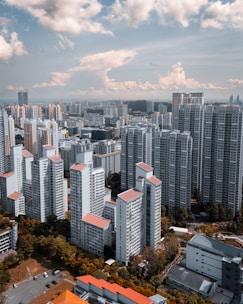 A spacious urban landscape is filled with high-rise apartment buildings and skyscrapers. The cityscape extends into the distance under a blue sky with scattered clouds. The buildings have a uniform architectural style with a dominant use of white and touches of red and orange on the rooftops. Lush greenery interspersed among the structures adds contrast to the scene.