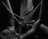 A person is intricately bound with ropes, creating a complex pattern around their body. The monochrome setting adds a stark contrast, emphasizing the textures of the skin and ropes. The person's eyes are closed, conveying a sense of calm or introspection.