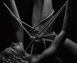 A person is intricately bound with ropes, creating a complex pattern around their body. The monochrome setting adds a stark contrast, emphasizing the textures of the skin and ropes. The person's eyes are closed, conveying a sense of calm or introspection.
