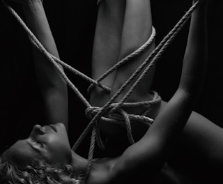 A person is intricately bound with ropes, creating a complex pattern around their body. The monochrome setting adds a stark contrast, emphasizing the textures of the skin and ropes. The person's eyes are closed, conveying a sense of calm or introspection.