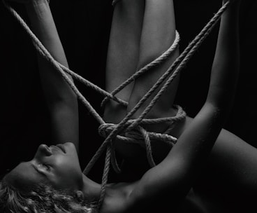 A person is intricately bound with ropes, creating a complex pattern around their body. The monochrome setting adds a stark contrast, emphasizing the textures of the skin and ropes. The person's eyes are closed, conveying a sense of calm or introspection.
