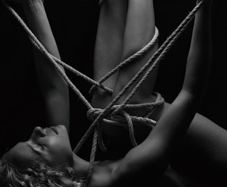 A person is intricately bound with ropes, creating a complex pattern around their body. The monochrome setting adds a stark contrast, emphasizing the textures of the skin and ropes. The person's eyes are closed, conveying a sense of calm or introspection.