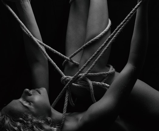 A person is intricately bound with ropes, creating a complex pattern around their body. The monochrome setting adds a stark contrast, emphasizing the textures of the skin and ropes. The person's eyes are closed, conveying a sense of calm or introspection.