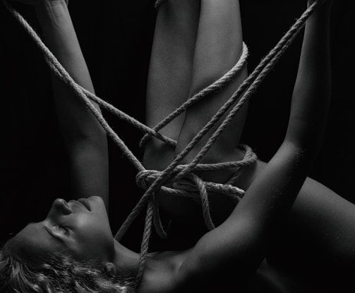A person is intricately bound with ropes, creating a complex pattern around their body. The monochrome setting adds a stark contrast, emphasizing the textures of the skin and ropes. The person's eyes are closed, conveying a sense of calm or introspection.